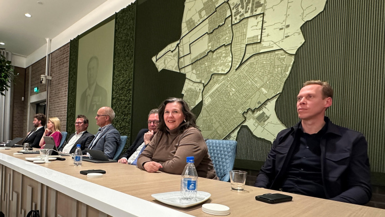 Mensen zitten aan een tafel in een vergaderruimte met een grote plattegrond aan de muur.