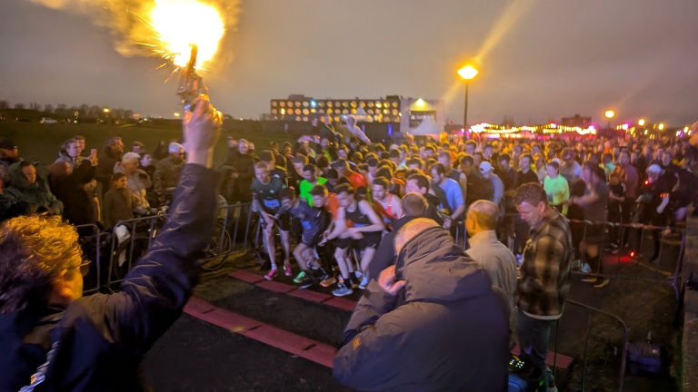 Een menigte hardlopers staat klaar voor de start van een nachtelijke race, aangemoedigd door toeschouwers en begeleid door een startschot.