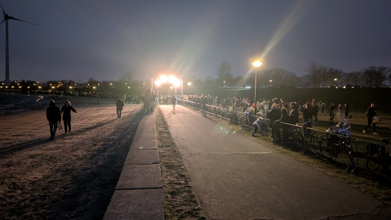 Nachtelijke scène met mensen op een verlicht pad langs het water, omringd door toeschouwers en fietsen.