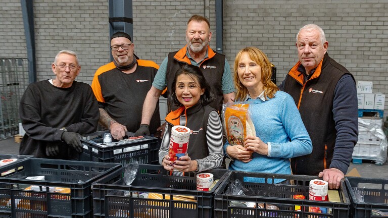 Een groep mensen poseert samen bij kratten met voedsel in een magazijnomgeving.