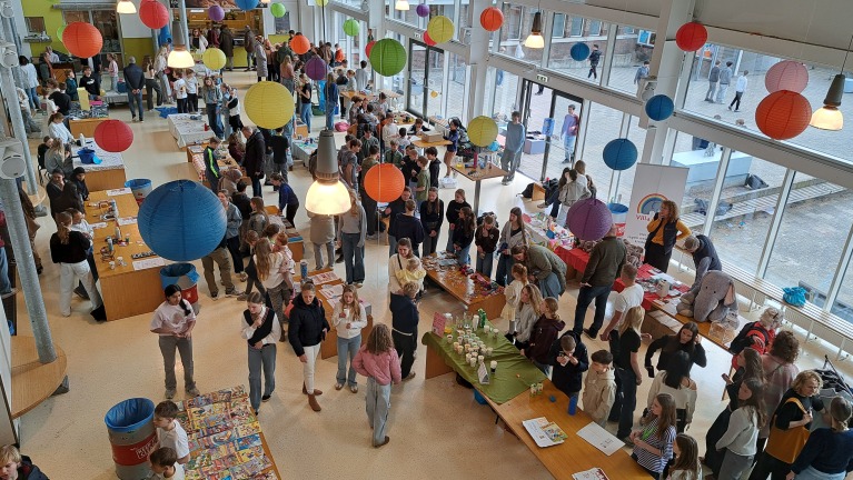 Een drukbezochte schoolbeurs met kleurrijke lampionnen, kraampjes en bezoekers, waaronder veel kinderen en volwassenen.