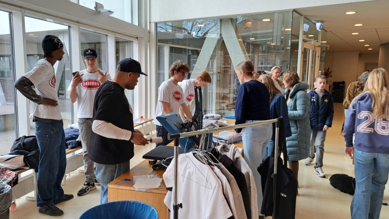 Mensen in een lichte ruimte met glazen wanden, bezig met kleding, sommige in shirts met 24SEVEN-logo.