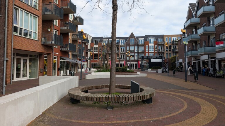 Modern plein met appartementsgebouwen en winkels, een boom met cirkelvormige bank in het midden, onder een bewolkte hemel.