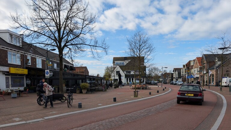 Straatbeeld met een rode auto op een bochtige weg, omgeven door winkels en cafés, onder een bewolkte blauwe hemel.
