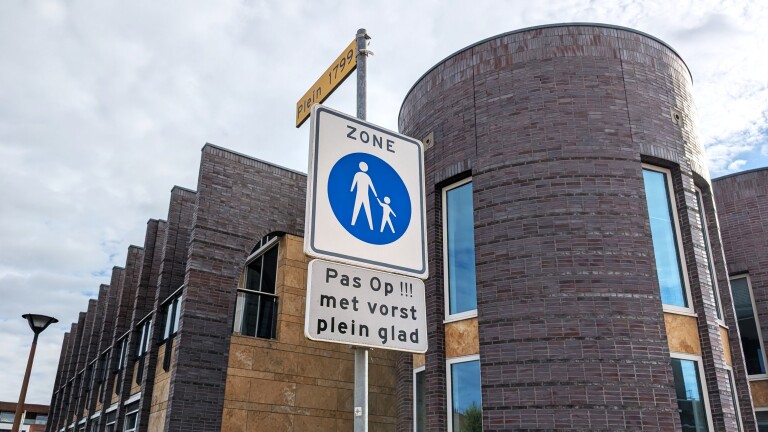 Verkeersbord met waarschuwing voor glad plein bij vorst, tegen de achtergrond van een modern, rond stenen gebouw.