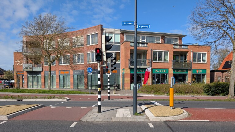 Bakstenen gebouw met winkels en kantoren, waaronder "day-one" en "IN-LOOP," gelegen bij een kruispunt met verkeerslichten en fietspad.