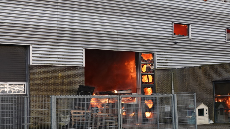 Brand in een industrieel gebouw met felle vlammen zichtbaar door ingeslagen ramen en een open deur, omheind terrein op de voorgrond.