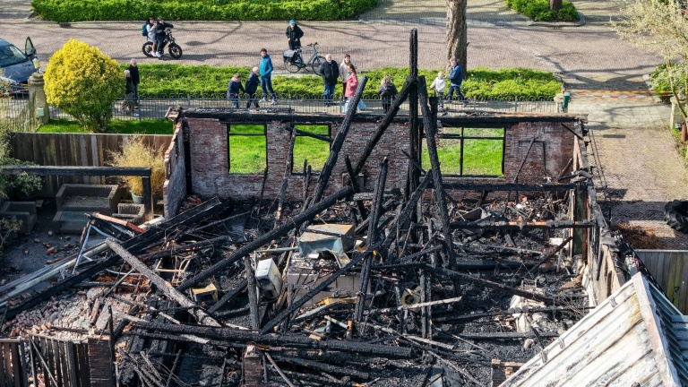 Verbrande resten van een huis, met zwartgeblakerde balken en puin; mensen kijken vanaf de straat toe.