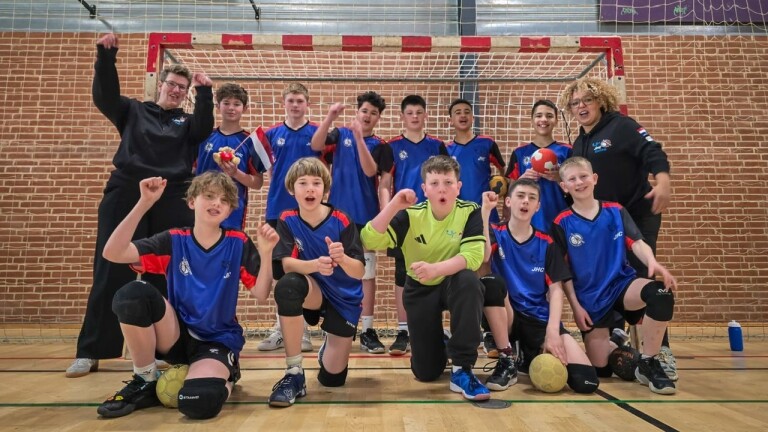 Een jeugdhandbalteam poseert enthousiast voor een doel met twee coaches, gekleed in blauwe sporttenues.