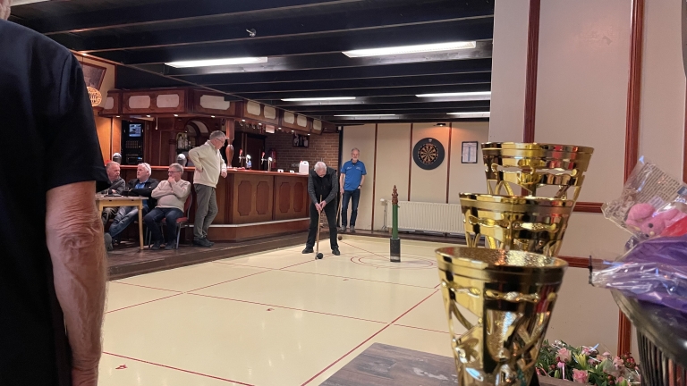 Een man speelt curling in een zaal met toeschouwers en trofeeën op de voorgrond.