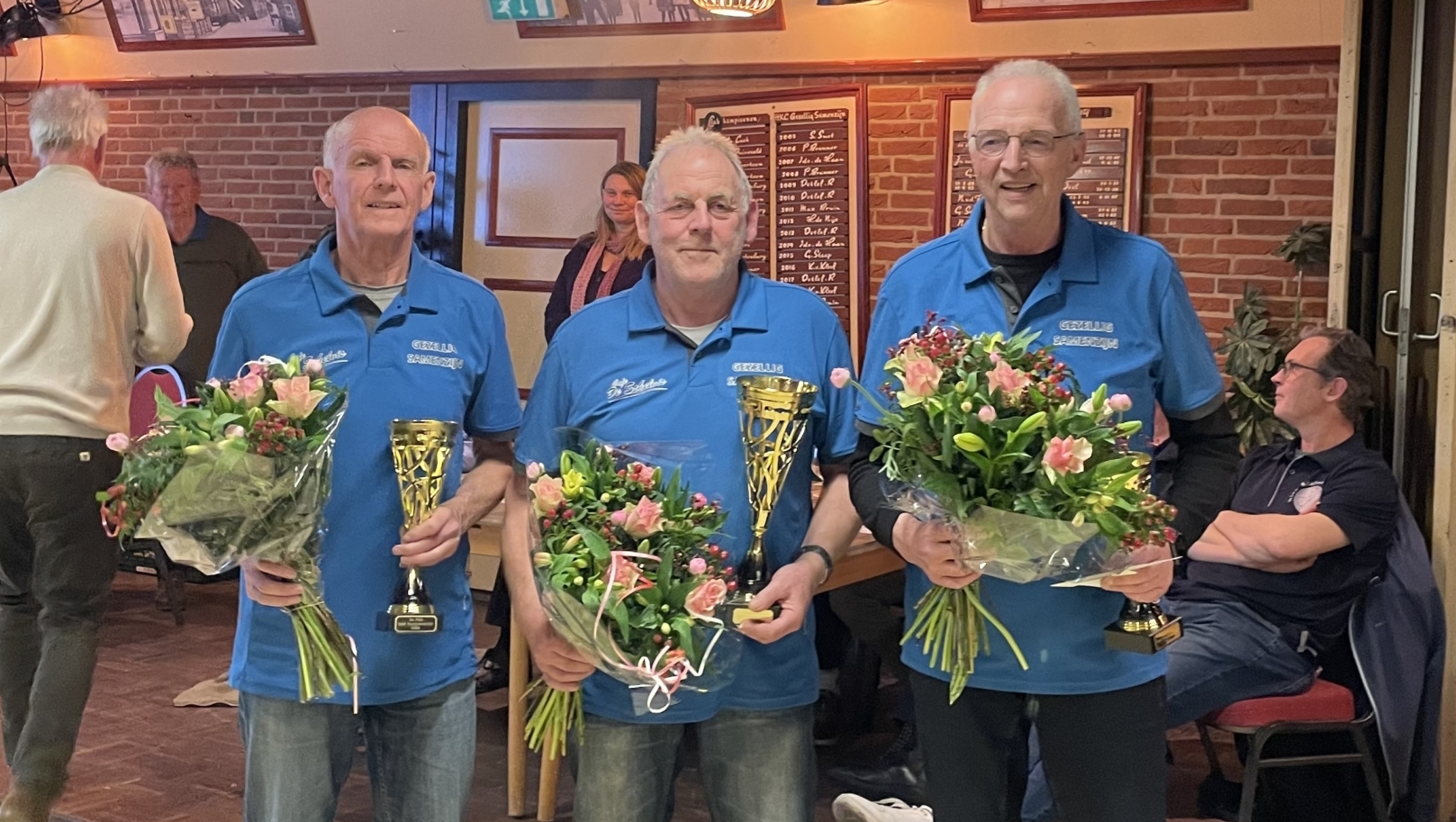 Drie mannen in blauwe shirts tonen trofeeën en boeketten in een zaal met andere mensen op de achtergrond.