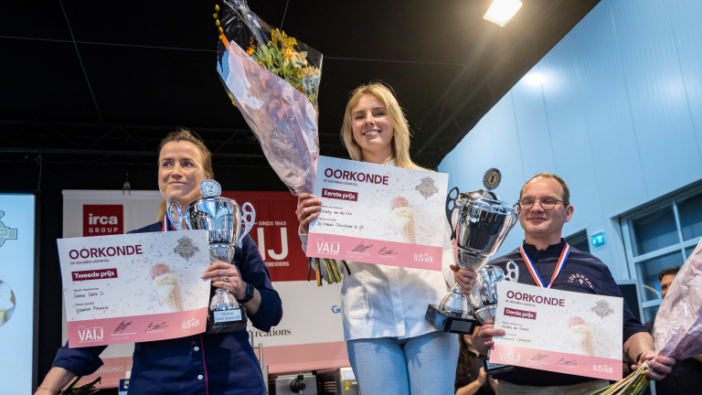 Drie mensen poseren met trofeeën, certificaten en bloemen na een prijsuitreiking.