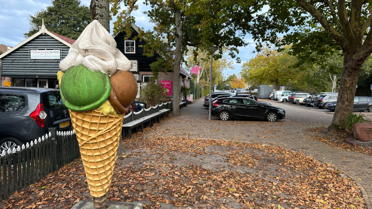 Groot ijsbeeld op een stoep omringd door herfstbladeren, met een fietsenwinkel op de achtergrond.