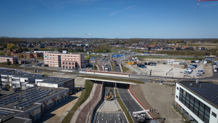 Luchtfoto van een bouwterrein met wegen en gebouwen, omgeven door een dorp en open landschap onder een heldere hemel.