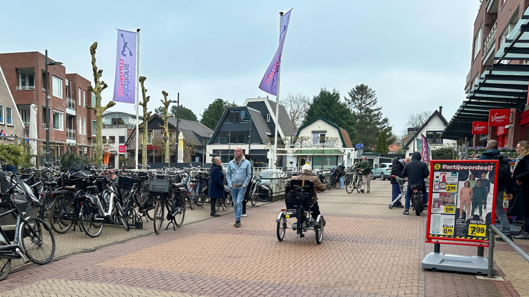 Straatbeeld met geparkeerde fietsen, enkele voetgangers en mensen voor een supermarkt met een reclamebord.