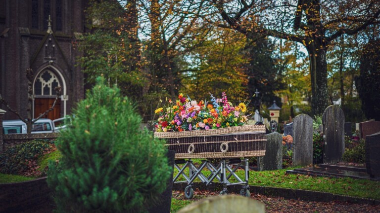 Een kist bedekt met kleurrijke bloemen staat op een baar in een kerkhof, omgeven door grafstenen en herfstbomen.