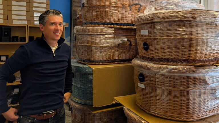 Een man staat naast stapels ingepakte, grote rotan manden in een magazijnachtige omgeving.