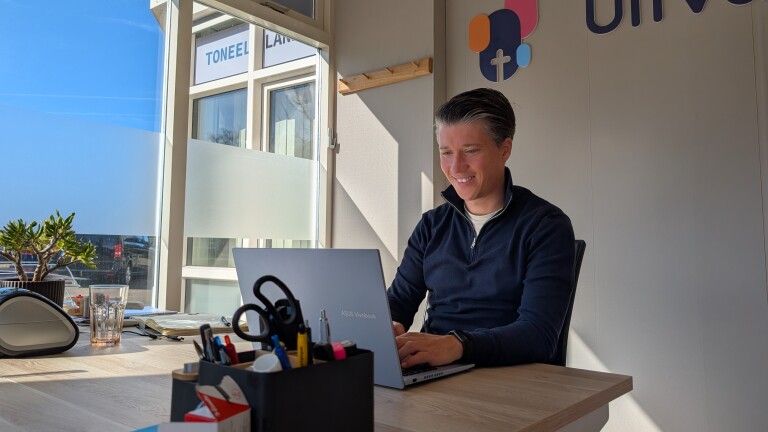 Persoon werkt glimlachend op een laptop aan een bureautafel met kantoorbenodigdheden en een plant.