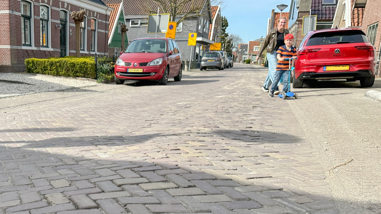 Kind op een step met een volwassene op een rustige straat met geparkeerde auto's en bakstenen huizen.