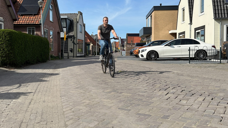 Man fietst in een zonnige straat met bakstenen huizen en geparkeerde auto's op de achtergrond.