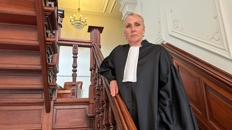 Een persoon in een toga staat op een trap van hout in een statige hal met een kroonluchter.