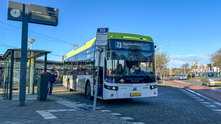 Bus 73 naar Haarlem overdag bij een bushalte, met een busstation in de achtergrond en een voorbijganger.