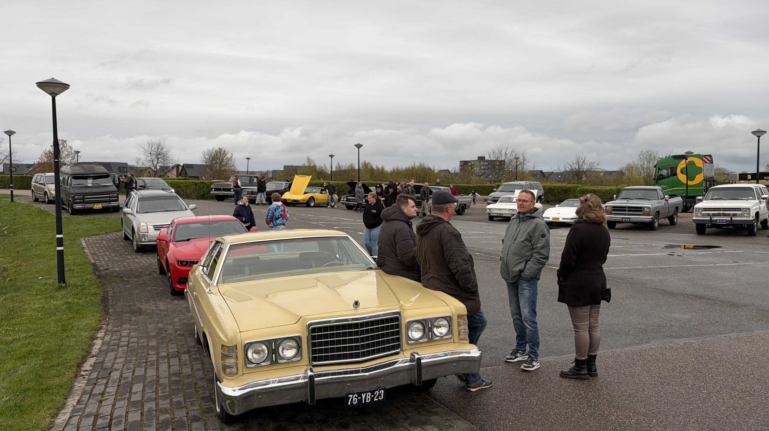 Groep mensen verzameld rond klassieke auto's op een parkeerplaats, met bewolkte lucht op de achtergrond.
