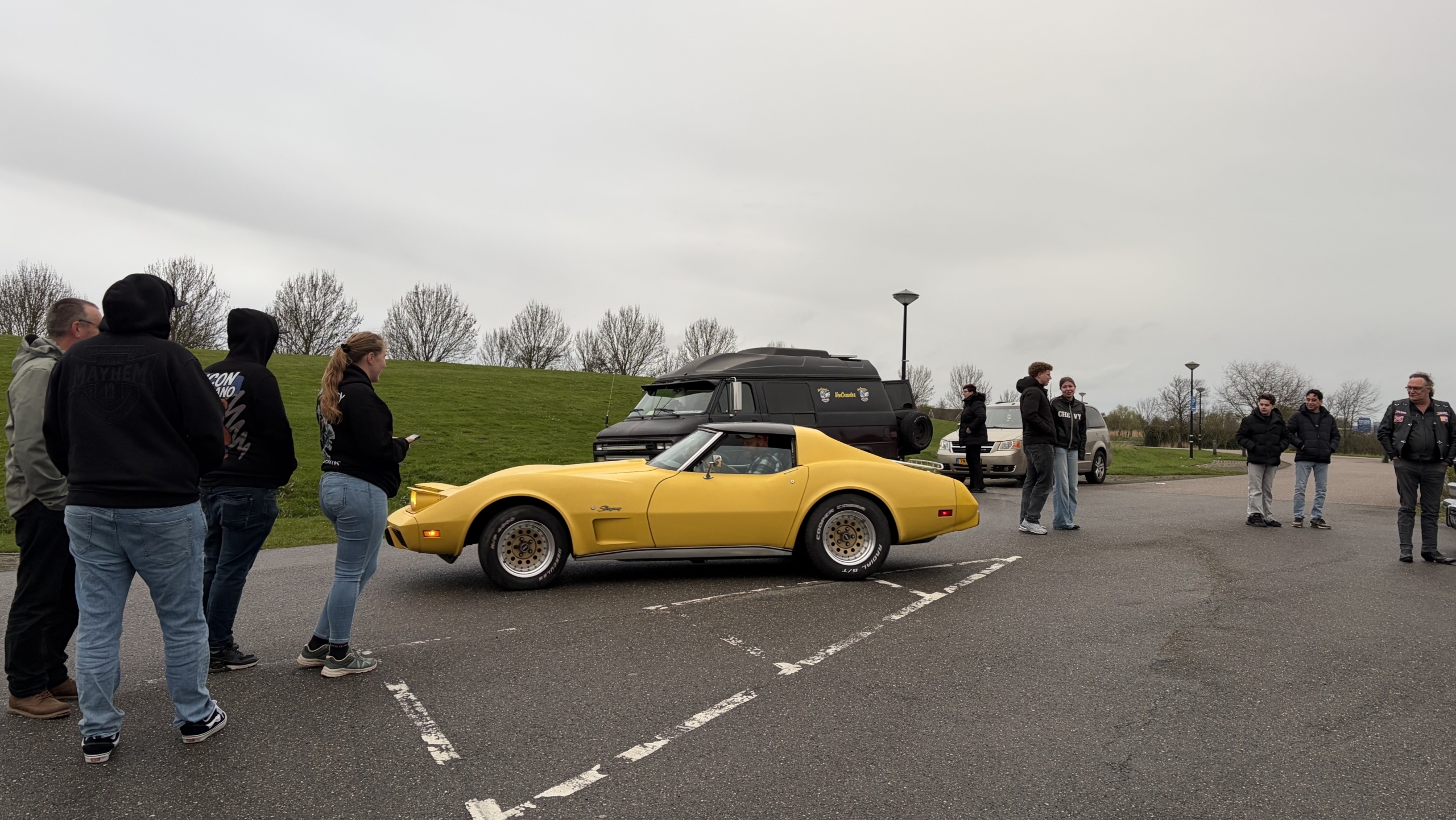 Een gele sportauto staat geparkeerd, omringd door mensen op een parkeerplaats met enkele bomen op de achtergrond.