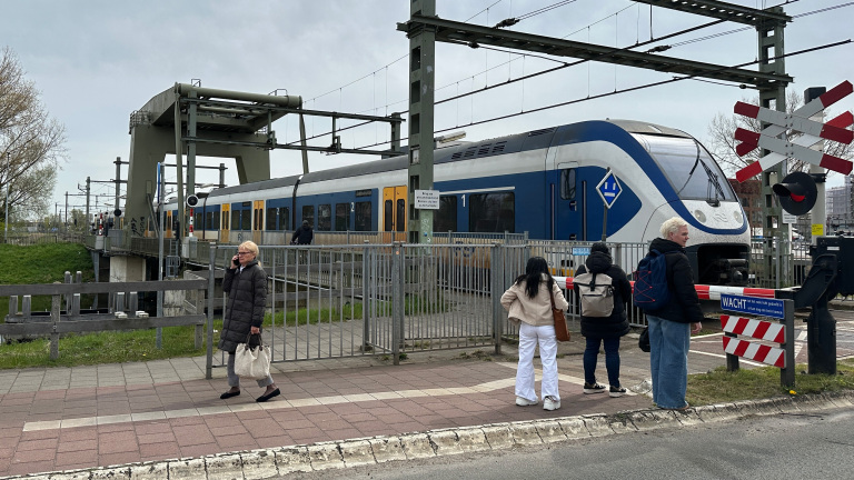 Een trein passeert een spoorwegovergang terwijl mensen op het trottoir wachten.
