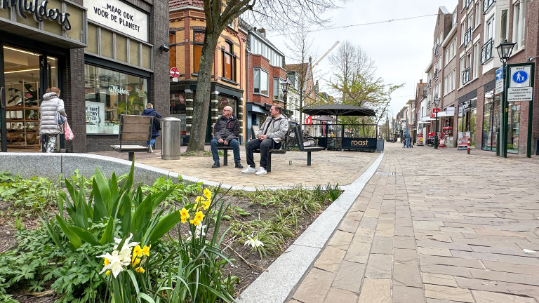 Mensen zitten op een bankje in een winkelstraat met bloeiende narcissen op de voorgrond.