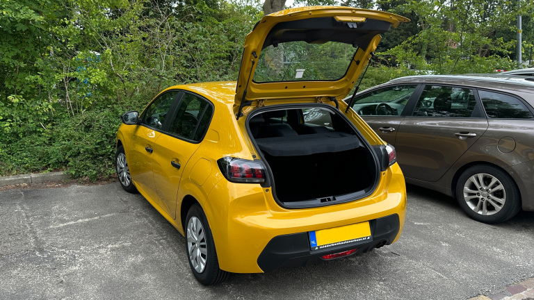 Gele auto met geopende achterklep geparkeerd naast een bruine auto, omgeven door groene struiken.