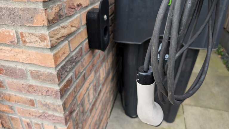 Een elektrische laadkabel hangt naast een bakstenen muur, met een laadstation op de achtergrond.