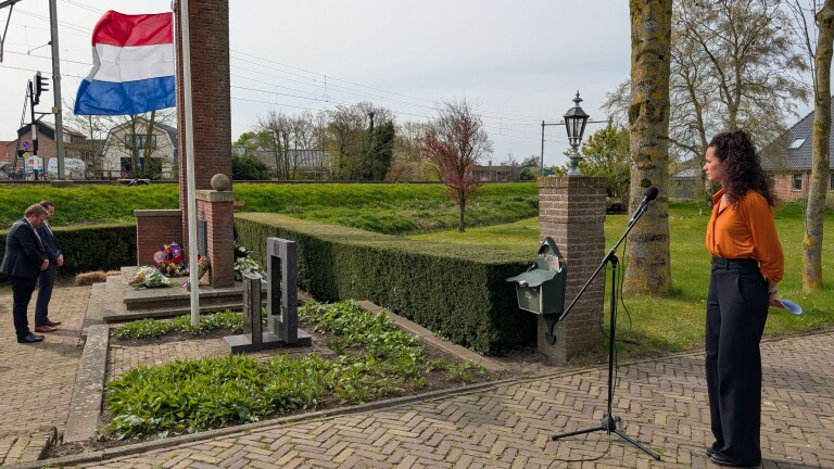 Twee mannen buigen hun hoofd bij een monument met bloemen en een Nederlandse vlag, terwijl een vrouw bij een microfoon staat.
