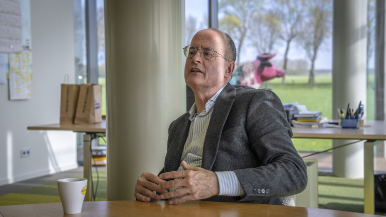 Man met bril en grijs jasje zit aan een tafel in een kantoor met papieren tasjes en een kartonnen beker.