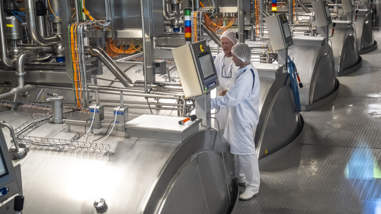 Twee mensen in witte beschermende kleding bedienen een monitor in een industriële fabriek met metalen apparaten en buizen.