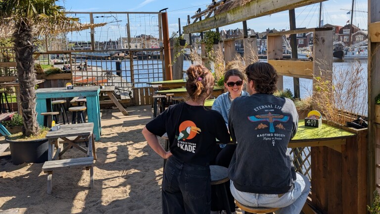 Drie mensen zitten aan een tafel in een strandbar nabij een haven, omgeven door houten meubels en planten.