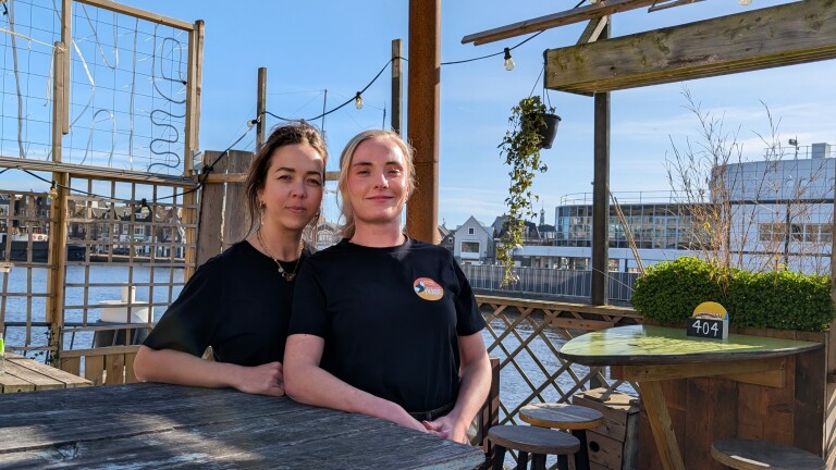 Twee vrouwen in zwarte shirts bij een houten tafel op een terras met uitzicht op een rivier en gebouwen op de achtergrond.