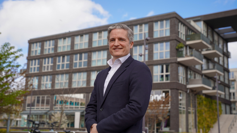Man in een donker pak staat buiten voor een modern gebouw, onder een blauwe lucht met enkele wolken.