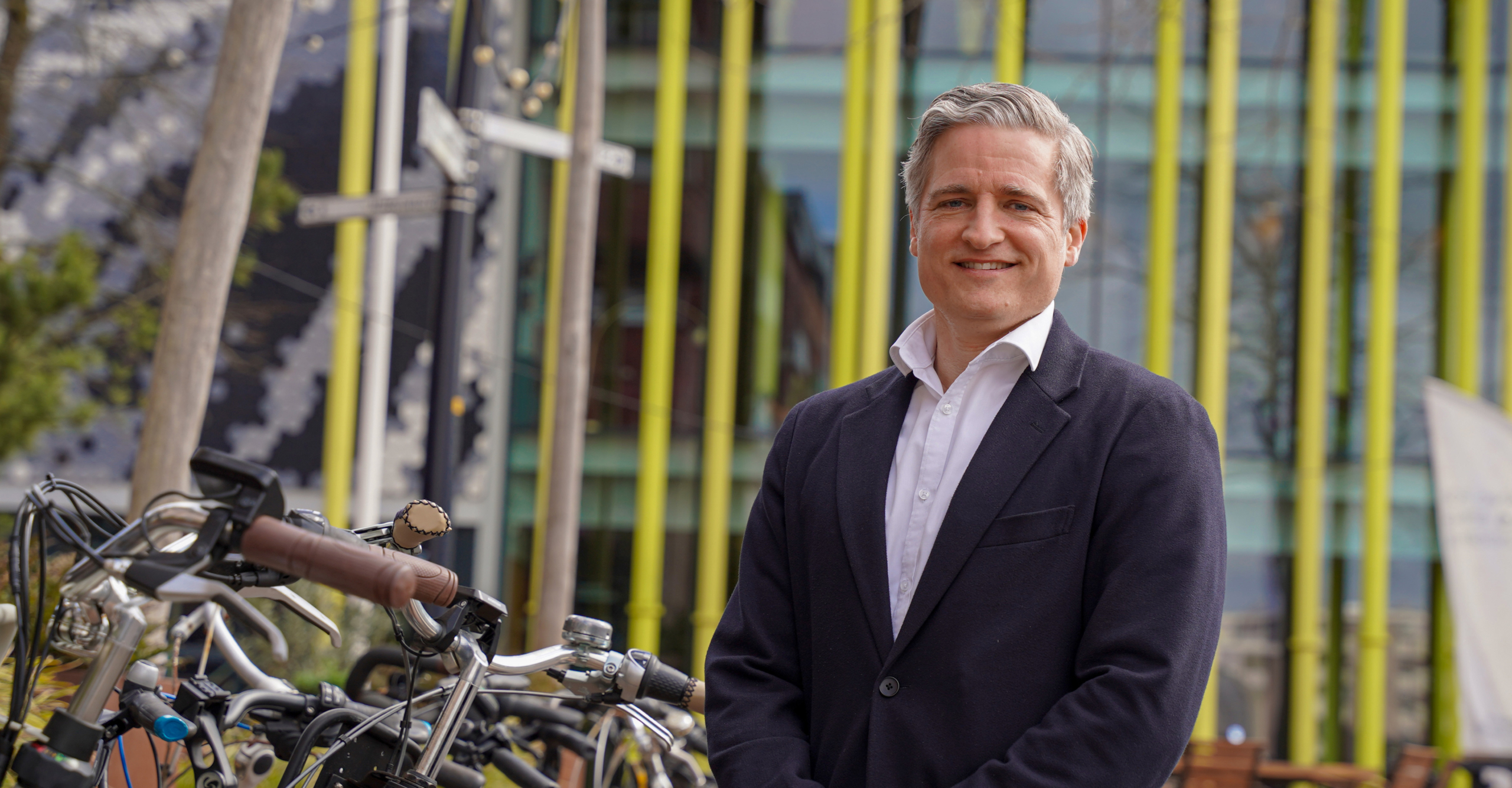 Man in een donker pak poseert naast fietsen, met een moderne gevel op de achtergrond.