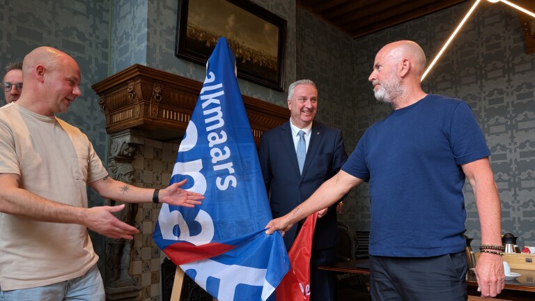 Drie mannen staan in een kamer rond een blauwe vlag met de tekst "Alkmaars". Een van hen draagt een pak.