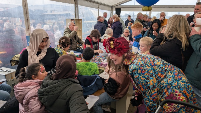 Mensen van verschillende leeftijden praten en vermaken zich in een drukke tent, met een vrouw in kleurrijke kleding en bloemenhoed op de voorgrond.