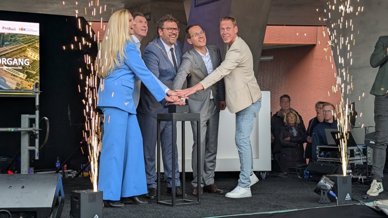 Een groep mensen in formele kleding houdt gezamenlijk een knop ingedrukt tijdens een ceremonieel moment met vonken op de achtergrond.
