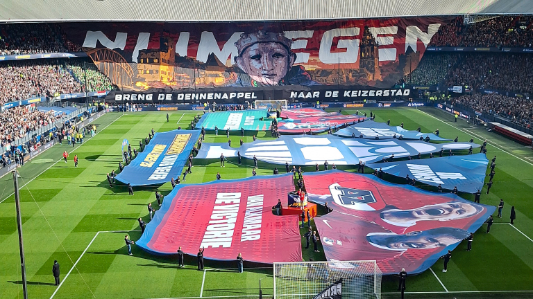 Voetbalstadion met groot spandoek in publiek met de tekst "Nijmegen" en grote doeken op het veld gedragen door mensen.