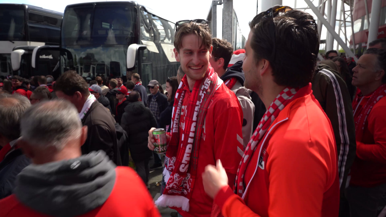 Twee mannen in rode sportkleding en sjaals staan lachend te praten in een menigte voor een rij bussen.