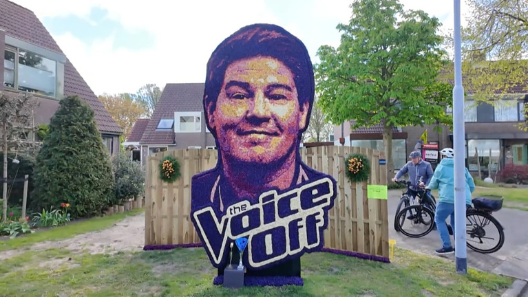 Bloemenmozaïek toont een gezicht en de tekst "the Voice off" op een hek, met fietsers en huizen op de achtergrond.