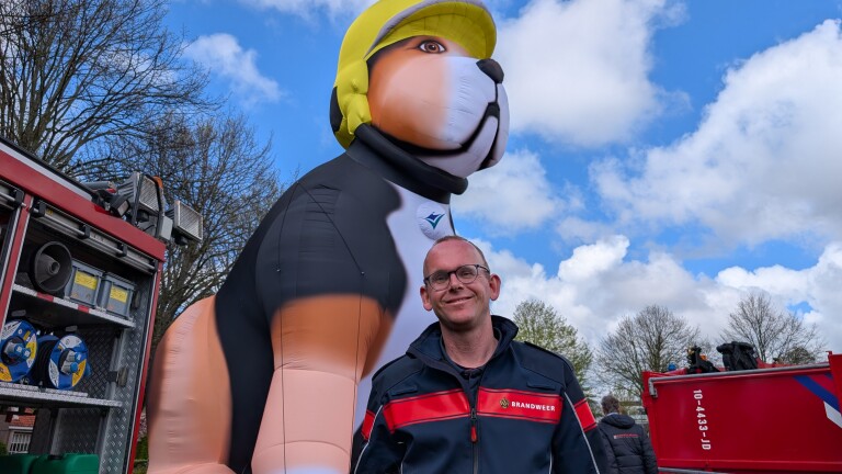 Man in brandweeruniform naast een grote opblaashond met brandweerhelmpje, bij een brandweerwagen onder een blauwe lucht.