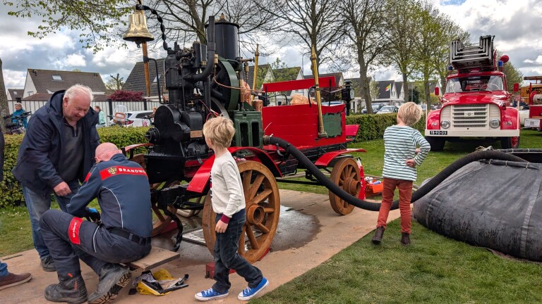 Kinderen en volwassenen bekijken een oude, rode stoombrandspuit naast een klassieke brandweerauto buiten.
