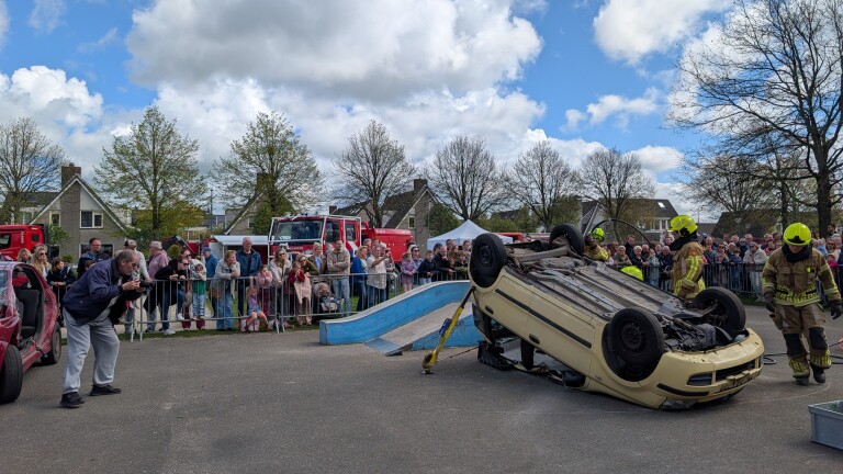 Brandweermensen demonstreren een auto-ongelukssituatie met een omgekeerde auto terwijl een menigte toekijkt.