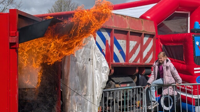 Een brandende container met vlammen, ernaast een meisje dat over een hekje bij een rode springkussen staat.