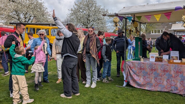 Mensen genieten van een drukbezochte markt met kraampjes, kleurrijke vlaggetjes en bloeiende bomen op de achtergrond.